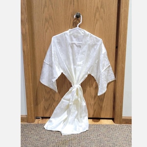 May include: A white, floral-patterned robe hangs on a white hanger from a black hook on a wooden door. The robe is tied at the waist with a matching sash. The fabric appears to be satin or a similar material.