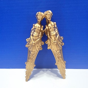 May include: A pair of gold-tone metal decorative accents featuring a stylized female figure design. The accents have a detailed, ornate design and a slightly curved shape.