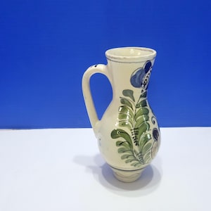 May include: A cream-colored ceramic pitcher featuring a hand-painted design of green leafy foliage and blue floral accents. The vessel has a curved handle on the side, a flared rim, and a rounded body that tapers down to a narrow base.