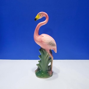 May include: A pink ceramic flamingo figurine with a yellow beak and green base. The flamingo is standing on a green leafy base. The figurine is a decorative item.
