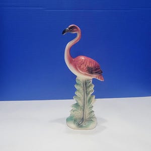 Puede incluir: Figura de flamenco de cerámica en tonos rosa, negro y blanco. El flamenco se alza sobre una base decorativa con un diseño de hojas. El ave tiene un cuello curvado y un pico negro.