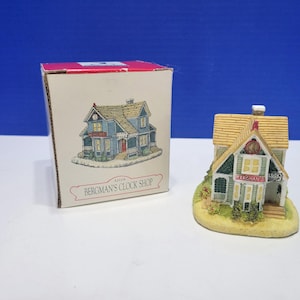 Può includere: Una statuetta in miniatura di un edificio intitolata Bergman's Clock Shop, caratterizzata da un esterno verde chiaro, un tetto color sabbia e un piccolo camino. È accompagnata dalla scatola originale in cartone bianco con l'illustrazione.