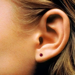 May include: A close-up of a person's ear with a small, round, red gemstone stud earring.