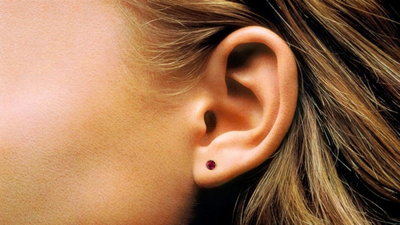 May include: A close-up of a person's ear with a small, round, red gemstone stud earring.