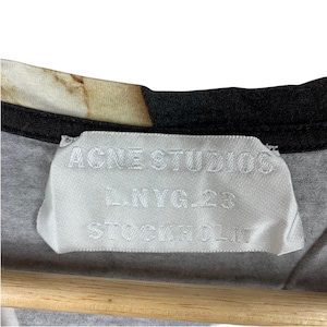 May include: A white fabric clothing label sewn into the inner neckline of a garment. The label features the text ACNE STUDIOS, L.NYG.23, and STOCKHOLM printed in a dark color. The garment has a black trim along the collar and a patterned fabric.