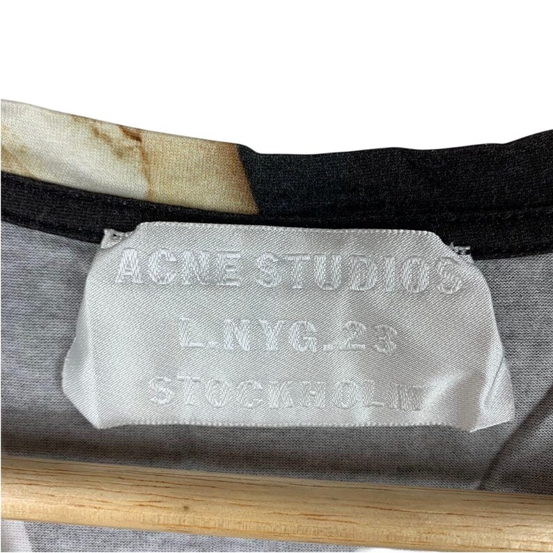 May include: A white fabric clothing label sewn into the inner neckline of a garment. The label features the text ACNE STUDIOS, L.NYG.23, and STOCKHOLM printed in a dark color. The garment has a black trim along the collar and a patterned fabric.