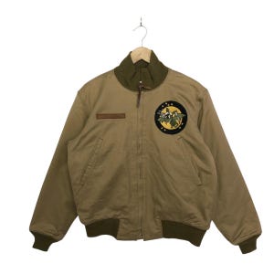 Può includere: Giacca bomber beige con colletto, polsini e fascia in vita verdi. La giacca presenta una toppa circolare e una toppa in pelle marrone sul petto. La giacca è appesa a una gruccia di metallo.