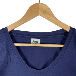 May include: A solid navy blue t-shirt with a scoop neckline. The garment features a white interior neck label with the text Acne printed in dark blue. The shirt is displayed on a light-coloured wooden hanger against a plain white background.