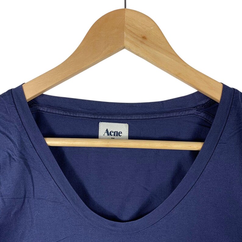 May include: A solid navy blue t-shirt with a scoop neckline. The garment features a white interior neck label with the text Acne printed in dark blue. The shirt is displayed on a light-coloured wooden hanger against a plain white background.