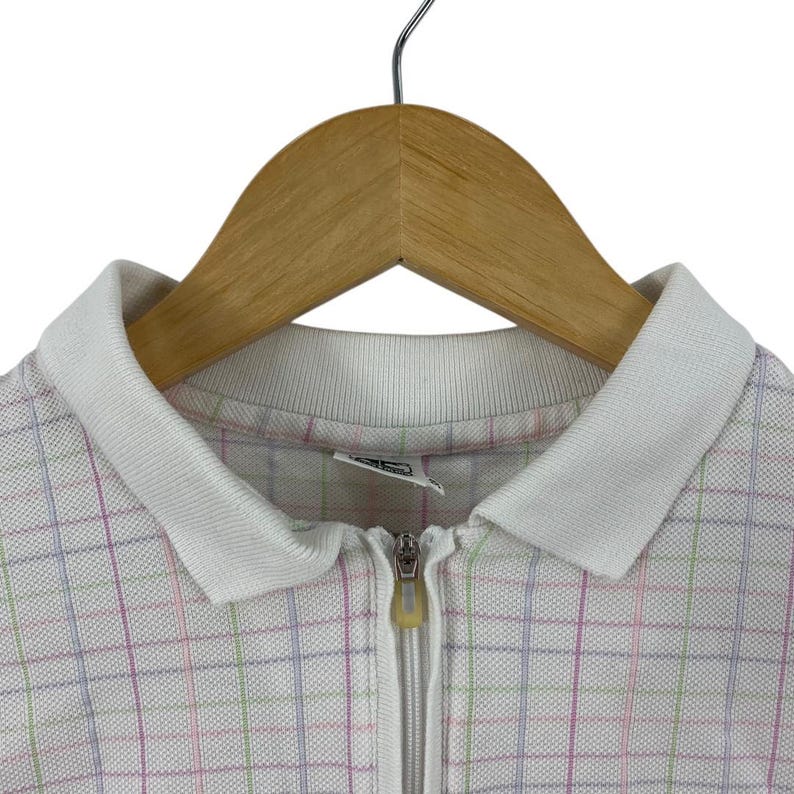 May include: A white zip-up polo shirt with a grid pattern in pastel pink, green, and blue. The shirt has a white collar and a full-length zipper. The garment is hanging on a wooden hanger.
