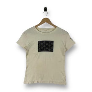 Può includere: Una t-shirt a maniche corte color crema con una toppa rettangolare nera sul petto caratterizzata da un motivo ripetuto del logo MCM. Un piccolo logo MCM secondario è stampato in nero sul bordo della manica destra. Girocollo classico.