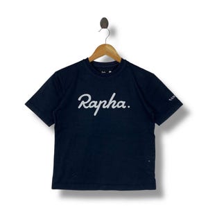 May include: Navy blue short-sleeve t-shirt with the white cursive text "Rapha" across the chest. The shirt is hanging on a wooden hanger. The brand name is also printed on the sleeve.
