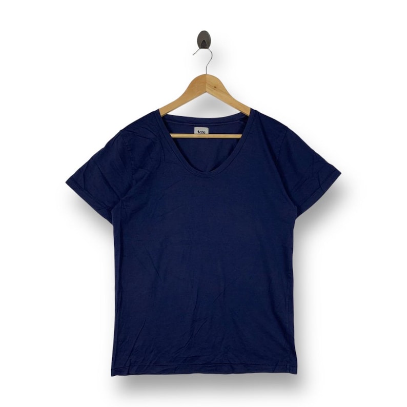 May include: A solid navy blue short-sleeve t-shirt with a scoop neckline, hanging on a wooden hanger. The inner collar features a white tag with the text Acne printed in black. The fabric appears to be a smooth cotton knit material.