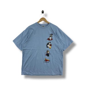 Könnte beinhalten: Hellblaues T-Shirt mit einem Cartoon-Motiv von Pinguinen in verschiedenen Posen. Das Shirt hat einen Rundhalsausschnitt und kurze Ärmel. Der Text "4 ELEMENTS" ist am unteren Rand des Motivs aufgedruckt.