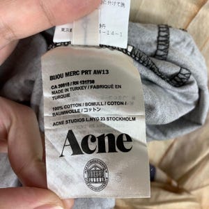 May include: A white fabric garment care label attached to a grey cotton textile. The label features black text including BIJOU MERC PRT AW13, CA 39815, RN 131738, MADE IN TURKEY, 100% COTTON, ACNE STUDIOS L.NYG.23 STOCKHOLM, the brand name Acne, and a logo.