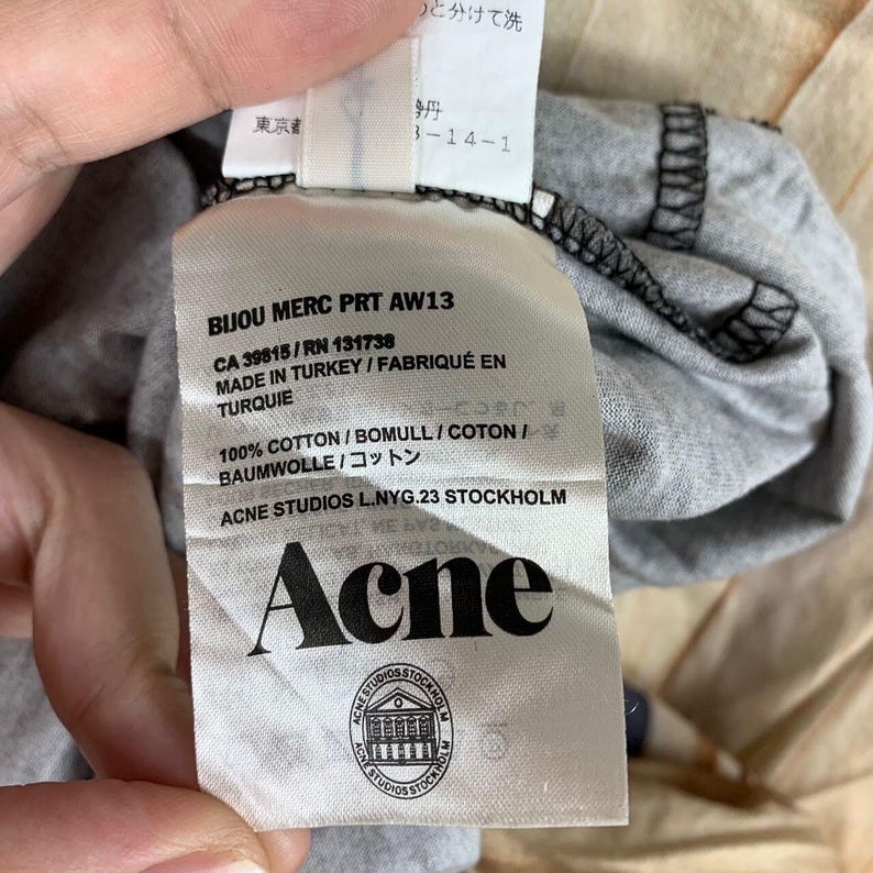 May include: A white fabric garment care label attached to a grey cotton textile. The label features black text including BIJOU MERC PRT AW13, CA 39815, RN 131738, MADE IN TURKEY, 100% COTTON, ACNE STUDIOS L.NYG.23 STOCKHOLM, the brand name Acne, and a logo.