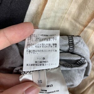 May include: A white fabric garment care label attached to a grey textile item. The label features Japanese text regarding washing instructions, followed by the text "トルコ製", "(株)三越伊勢丹", and "東京都新宿区新宿 3-14-1". A smaller tag below displays the size "XS" and "W13", with "KEY / FABRIQU&Eacute; EN" visible.