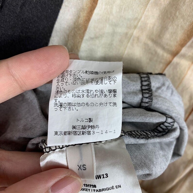 May include: A white fabric garment care label attached to a grey textile item. The label features Japanese text regarding washing instructions, followed by the text "トルコ製", "(株)三越伊勢丹", and "東京都新宿区新宿 3-14-1". A smaller tag below displays the size "XS" and "W13", with "KEY / FABRIQU&Eacute; EN" visible.