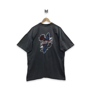 Op de afbeelding: Donkergrijs T-shirt met een afbeelding op de achterkant met een blauw surfboard, een rode roos en de woorden "Killer Love" op een banner. Het T-shirt hangt aan een houten hanger.