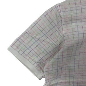 May include: Close-up of a white polo shirt sleeve with a colorful plaid pattern. The plaid features thin lines of pink, green, and blue, creating a subtle, textured design. The sleeve has a white ribbed cuff.