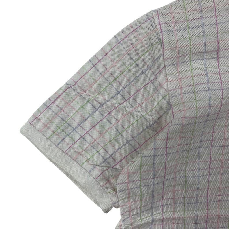 May include: Close-up of a white polo shirt sleeve with a colorful plaid pattern. The plaid features thin lines of pink, green, and blue, creating a subtle, textured design. The sleeve has a white ribbed cuff.