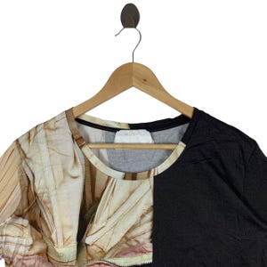 May include: A short-sleeve t-shirt featuring a split design with a solid black panel on the right side and a photographic print of draped, light-colored fabric on the left side. The shirt has a round neckline and is displayed on a wooden hanger.