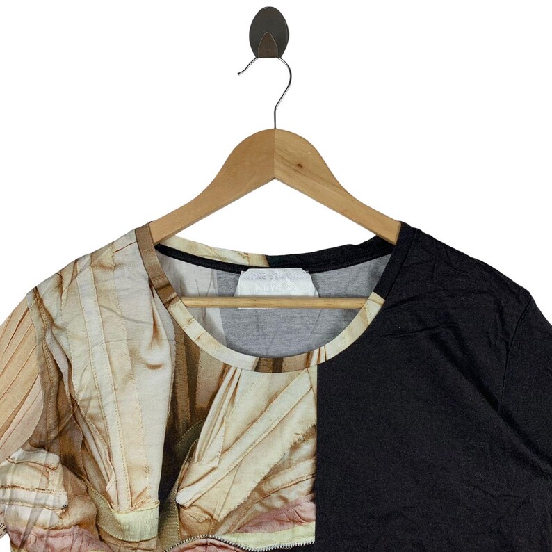 May include: A short-sleeve t-shirt featuring a split design with a solid black panel on the right side and a photographic print of draped, light-colored fabric on the left side. The shirt has a round neckline and is displayed on a wooden hanger.