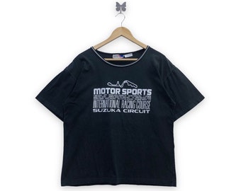 Può includere: T-shirt nero con bordatura bianca. La maglietta presenta la scritta "MOTOR SPORTS INTERNATIONAL RACING COURSE SUZUKA CIRCUIT" in bianco. Una grafica di un circuito automobilistico è sopra il testo. La maglietta è appesa.