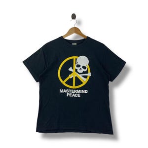 May include: Black t-shirt with a yellow peace sign and a white skull and crossbones graphic. The text "MASTERMIND PEACE" is printed below the graphic. The shirt is hanging on a wooden hanger.
