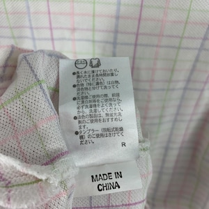 May include: Close-up of a white fabric with a plaid pattern in pink, green, and blue. A tag with Japanese text and the words "MADE IN CHINA" is attached to the fabric. The fabric appears to be part of a garment.