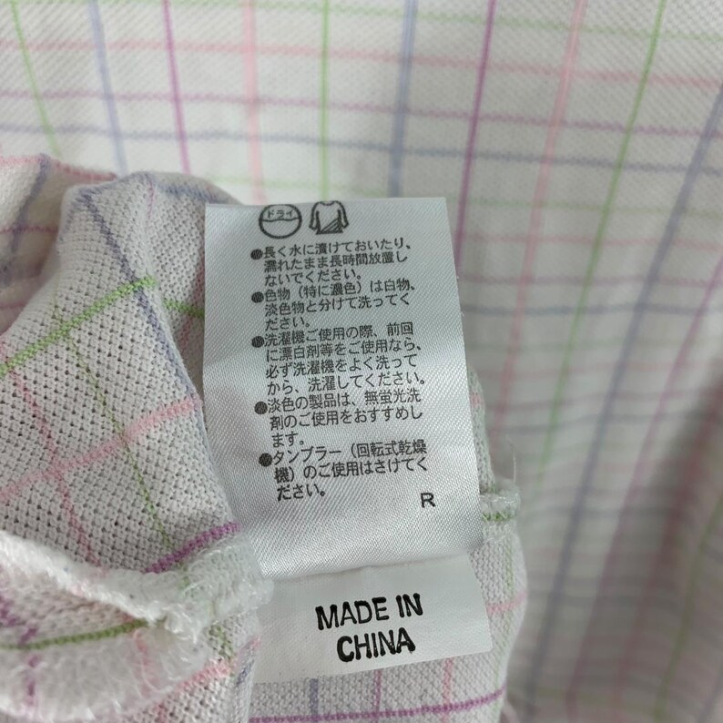 May include: Close-up of a white fabric with a plaid pattern in pink, green, and blue. A tag with Japanese text and the words "MADE IN CHINA" is attached to the fabric. The fabric appears to be part of a garment.