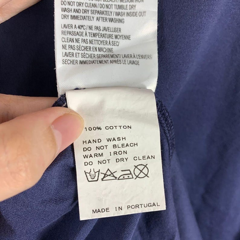 May include: A navy blue cotton garment featuring a white fabric care label with black text. The label specifies 100% cotton, hand wash, do not bleach, warm iron, and do not dry clean, with care symbols and the text Made in Portugal at the bottom.