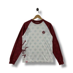 May include: White long-sleeved shirt with maroon sleeves and a crew neck. The shirt features a pattern of light blue floral designs and a graphic of a figure with long arms. A small emblem is on the chest.