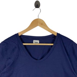 May include: A dark blue t-shirt with a scoop neckline, hanging on a wooden hanger. The shirt features a white interior neck label with the text Acne printed in black. The fabric appears to be a solid, smooth cotton material with short sleeves.