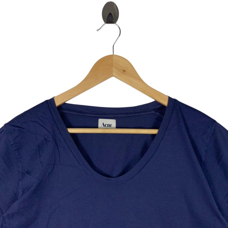 May include: A dark blue t-shirt with a scoop neckline, hanging on a wooden hanger. The shirt features a white interior neck label with the text Acne printed in black. The fabric appears to be a solid, smooth cotton material with short sleeves.