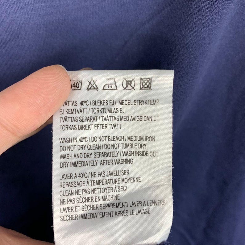 May include: A white rectangular garment care label with black text and laundry symbols sewn onto a dark blue fabric. The label includes instructions in Swedish, English, and French, specifying washing at 40 degrees Celsius and drying.