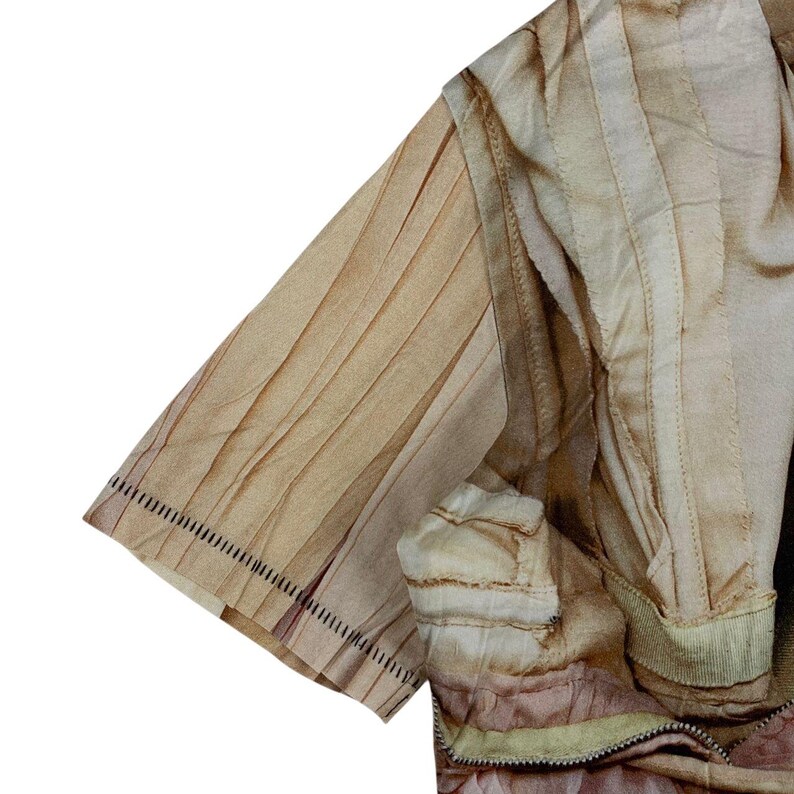 May include: A short-sleeved garment featuring a light beige and tan color palette with vertical pleated detailing. The fabric displays a subtle gradient effect, and the sleeve hem is finished with a distinct row of dark, visible stitching.