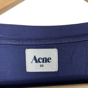 May include: A dark blue t-shirt featuring a rectangular white fabric label sewn into the inner neckline. The label displays the text Acne in a dark blue serif font, with the size XS printed in a smaller dark blue font directly underneath the brand name.