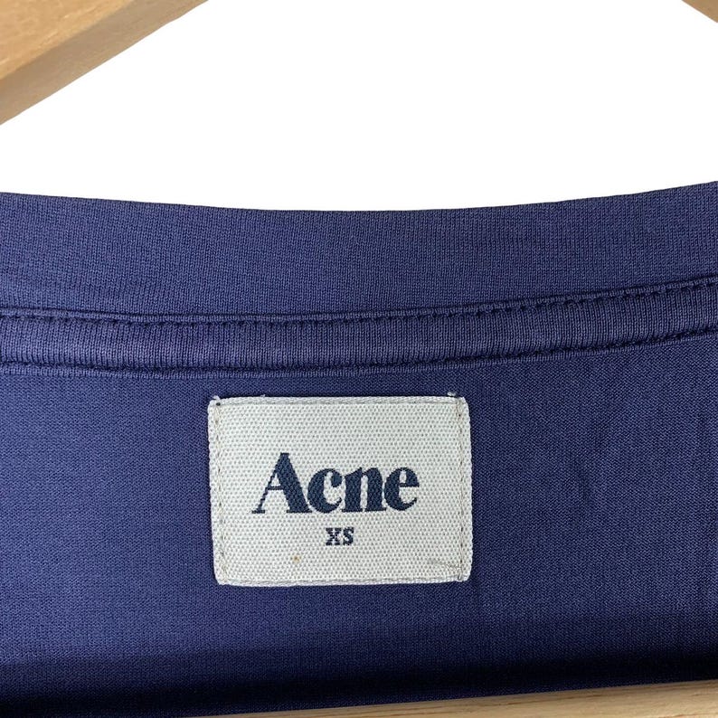 May include: A dark blue t-shirt featuring a rectangular white fabric label sewn into the inner neckline. The label displays the text Acne in a dark blue serif font, with the size XS printed in a smaller dark blue font directly underneath the brand name.