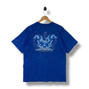May include: Royal blue t-shirt with a graphic design on the back. The design features a stylized wave pattern in shades of blue and white, with the text "Puto Hawaiian Longboard Wear". The shirt is hanging on a wooden hanger.