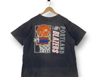 Puede incluir: Una camiseta de manga corta gris carbón con un gráfico de una canasta de baloncesto y el texto 1991 NBA PLAYOFFS. La prenda también muestra el texto PORTLAND BLAZERS en letras blancas y rojas junto al logotipo del equipo en el frente.