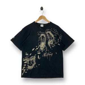 Peut inclure: T-shirt noir avec un motif beige représentant des créatures à gueules ouvertes et de nombreux yeux. Le mot "Hellhound" est écrit dans une police stylisée. Le t-shirt est suspendu à un cintre en bois.