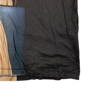 May include: A black t-shirt featuring a printed graphic on the left side. The graphic displays a vertical section of a beige fabric drape next to a dark blue rectangular area. The fabric has visible folds and a hemline along the bottom edge.