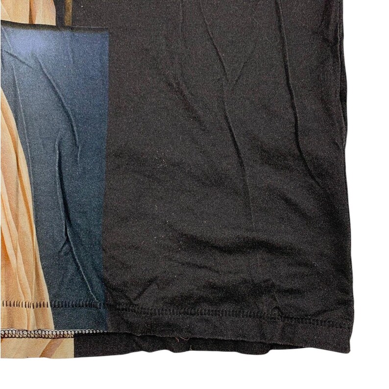 May include: A black t-shirt featuring a printed graphic on the left side. The graphic displays a vertical section of a beige fabric drape next to a dark blue rectangular area. The fabric has visible folds and a hemline along the bottom edge.