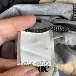 May include: A white fabric garment care label attached to a grey heathered textile. The label features the text 100% in Japanese characters for cotton, followed by a series of five laundry care symbols including a washing icon, a no-bleach icon, and a low-heat iron icon.