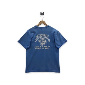 May include: A blue t-shirt with a vintage graphic on the back, featuring the text "J. Buegeleisen Co. Enthusiast" and "Buco Helmets." The shirt is hanging on a wooden hanger against a white background.