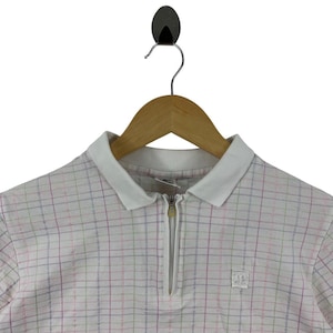 May include: White polo shirt with a colorful plaid pattern. The shirt has a white collar and a quarter-length zipper. A small logo is visible on the right side of the shirt. The shirt is hanging on a wooden hanger.