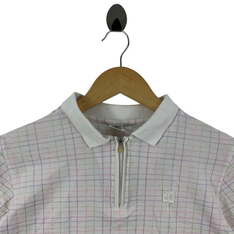 May include: White polo shirt with a colorful plaid pattern. The shirt has a white collar and a quarter-length zipper. A small logo is visible on the right side of the shirt. The shirt is hanging on a wooden hanger.