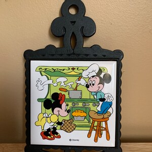 May include: A black cast iron hot pad with a ceramic tile featuring Mickey Mouse and Minnie Mouse cooking in a kitchen. The tile has a green background and features a yellow stove, a red pot, a brown pie, and a wooden stool. The tile is surrounded by a black cast iron frame with a decorative top.