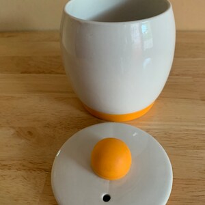 May include: White ceramic egg-shaped container with a yellow rim and a matching lid. The lid has a yellow knob on top.